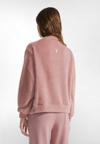 Pullover in felpa rosa soffice con colletto rotondo, vestibilità rilassata e orlo a coste. Presenta un piccolo tag con logo sul retro. Texture morbida.