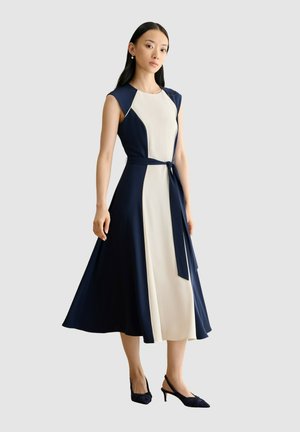 Frau trägt ein ärmelloses, marineblau-weißes Midikleid mit gebundenem Taillengürtel und schwarzen Slingback-Schuhen mit niedrigem Absatz, steht vor grauem Hintergrund.