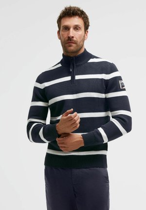 Man wearing a navy blue and white striped knit sweater with a quarter zipper, standing against a plain light background.