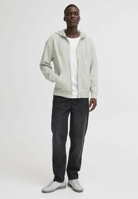 Felpa grigia chiara con zip e cordoncini, abbinata a una maglietta bianca e jeans scuri. Il modello indossa sneakers grigie. Sfondo neutro.