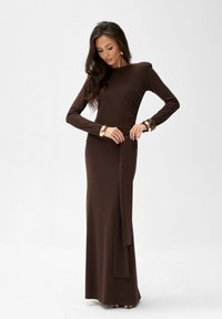 Brązowa sukienka z długim rękawem, wykonana z gładkiego, elastycznego materiału, z dopasowanym gorsetem i rozcięciem w spódnicy. Dodatki to złote bransoletki.