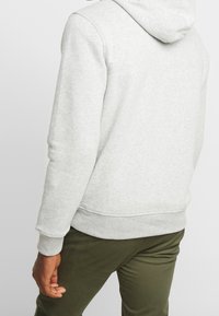 Hellgrauer Hoodie mit Kängurutasche, gerippten Bündchen und Saum. Das Material wirkt weich und strukturiert, mit einer Kapuze mit Kordelzug. Getragen mit dunkelgrünen Hosen.