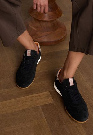 Black suede sneakers with a white sole, featuring a patterned heel and laces, placed on a wooden floor. Accented with a brown logo tag.