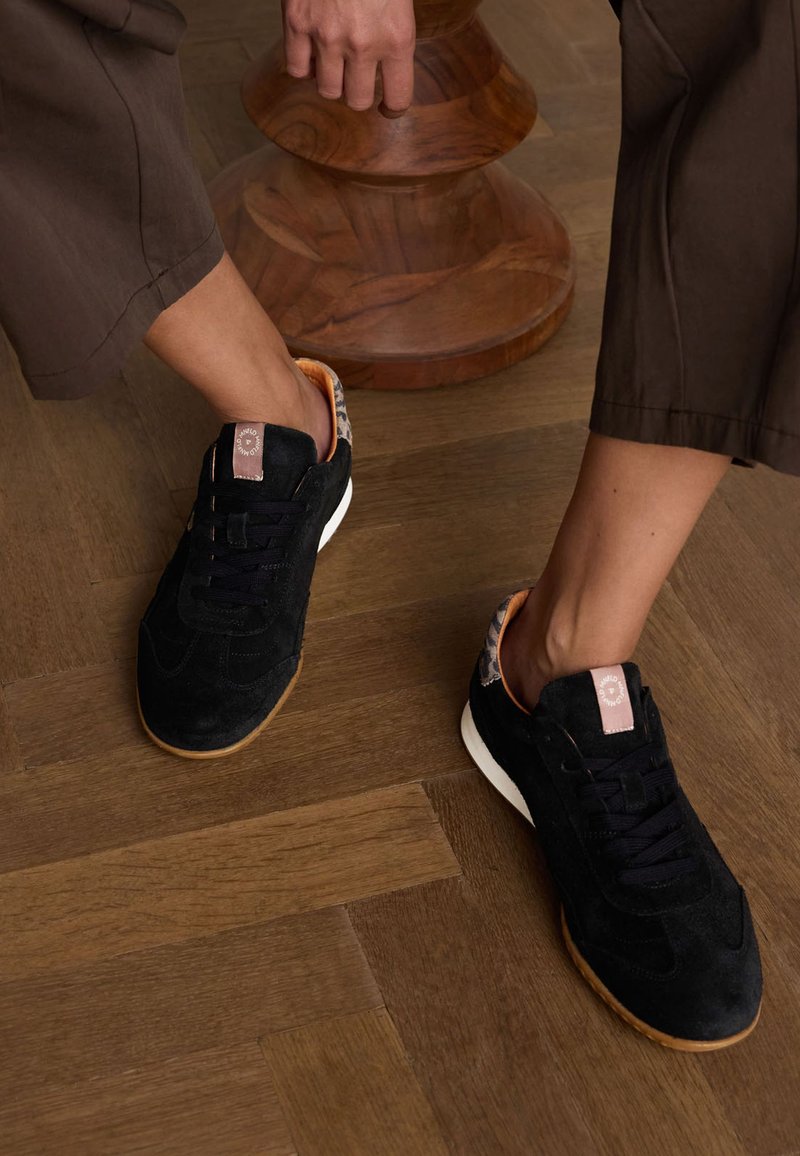 Sneakers in suede nera con suola bianca, caratterizzati da un tallone e lacci a fantasia, posati su un pavimento in legno. Accentuati da un'etichetta con logo marrone.