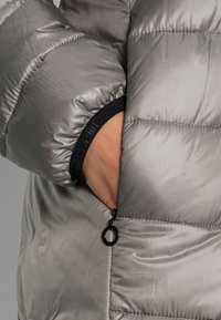 Ręka włożona do zasuwanej bocznej kieszeni błyszczącej szarej kurtki puchowej, z widocznym czarnym elastycznym mankietem.