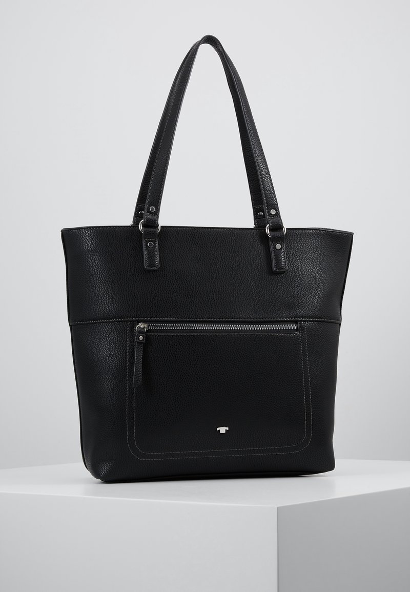 Sac cabas en cuir noir avec deux bretelles, poche avant zippée et logo discret, présenté sur un piédestal blanc sur fond gris.