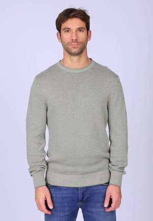 Homme portant un pull gris clair texturé et un jean bleu, debout devant un fond violet clair uni.