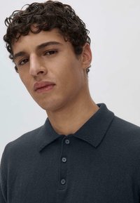 Dark gray polo shirt featuring a knit texture, a classic collar, and three button placket. Model has short, curly hair.