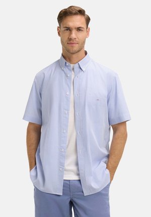 Homme portant une chemise bleu clair à manches courtes boutonnée sur un t-shirt blanc avec un pantalon bleu, les mains dans les poches, regardant devant lui avec une expression neutre.
