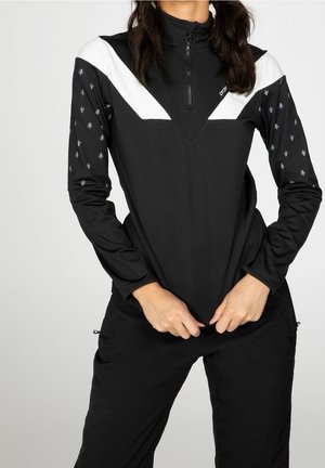 Persoon draagt een zwart sportshirt met witte chevron en kleine boompatronen op de mouwen, en past met beide handen de zoom aan.