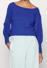 Modrý pletený svetřík se spadlými rameny, dlouhými nafouklými rukávy a žebrovanými manžetami, nošený s světlomodrými kalhotami širokých nohavic.