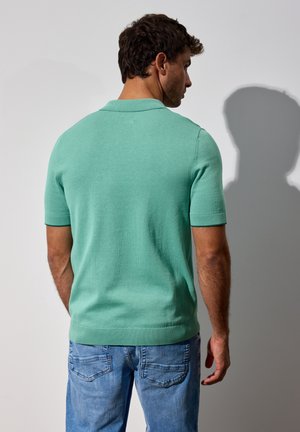 Kurzärmeliges Polohemd aus hellgrüner Baumwolle mit geripptem Kragen und Bündchen, kombiniert mit hellblauen Jeans. Rückansicht, minimalistische Details.