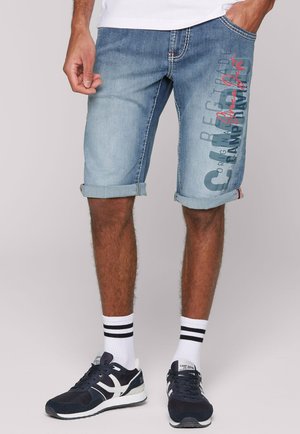 Hellblaue Denim-Shorts mit umgeschlagenem Saum, versehen mit auffälligen Textgrafiken auf der Vorderseite in dunkleren und roten Tönen. Kombiniert mit dunklen Sneakers.