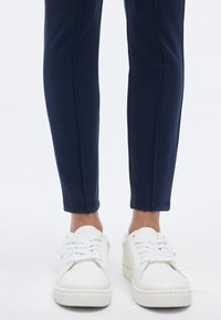 Λευκά αθλητικά παπούτσια με υφή στις καουτσούκ σόλες και σχέδιο με κορδόνια, συνδυασμένα με εφαρμοστά σκούρα μπλε παντελόνια με λείο ύφασμα και στενή γραμμή στα πόδια.