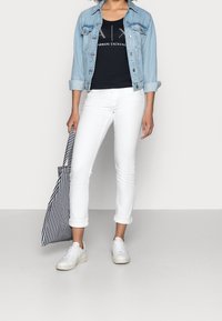 Světle modrá denimová bunda, černý top s logem, bílé slim džíny s vyhrnutými nohavicemi a bílé tenisky. Drží černobílou pruhovanou tašku.