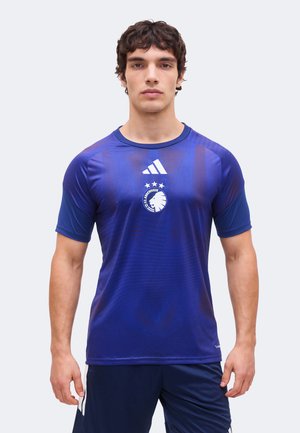 Mann trägt ein blaues Adidas-Sportshirt mit dem Logo von FC Kopenhagen und drei Sternen, steht vor einem schlichten hellen Hintergrund.