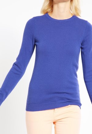 Femme portant un pull ajusté à manches longues bleu roi et un pantalon beige clair sur un fond blanc uni.