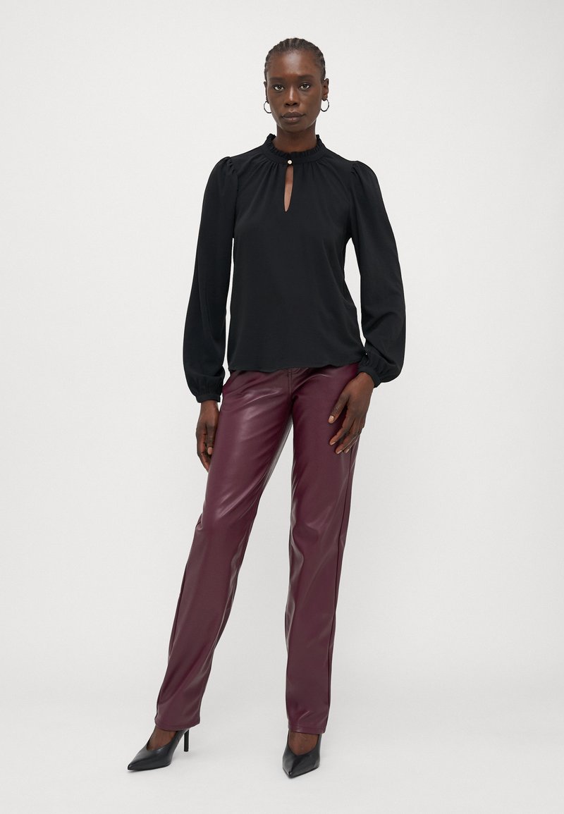 Blusa nera a maniche lunghe con dettaglio a occhiello sul collo, abbinata a pantaloni di pelle bordeaux e tacchi neri a punta. Tessuto liscio, vestibilità sartoriale.