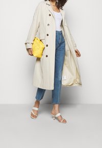 Gabardina beige con grandes botones, de tejido ligero, llevada sobre una camiseta blanca y jeans azules. Bolso de mano tejido amarillo y sandalias de tacón blancas.
