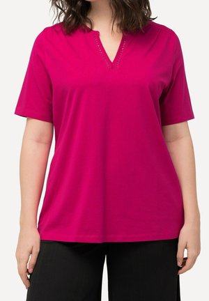 Femme portant un t-shirt rose vif à manches courtes avec un col en V et des détails cousus subtils, associée à un pantalon noir, debout sur un fond blanc.