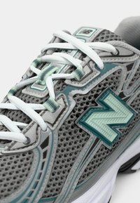 Gros plan sur une chaussure de sport grise et turquoise avec des lacets blancs, en tissu en maille et un logo "N" texturé sur le côté.
