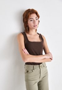 Smeđa majica bez rukava sa kvadratnim izrezom u kombinaciji s maslinasto zelenim trapericama visokog struka. Model stoji s prekriženim rukama uz neutralnu pozadinu.