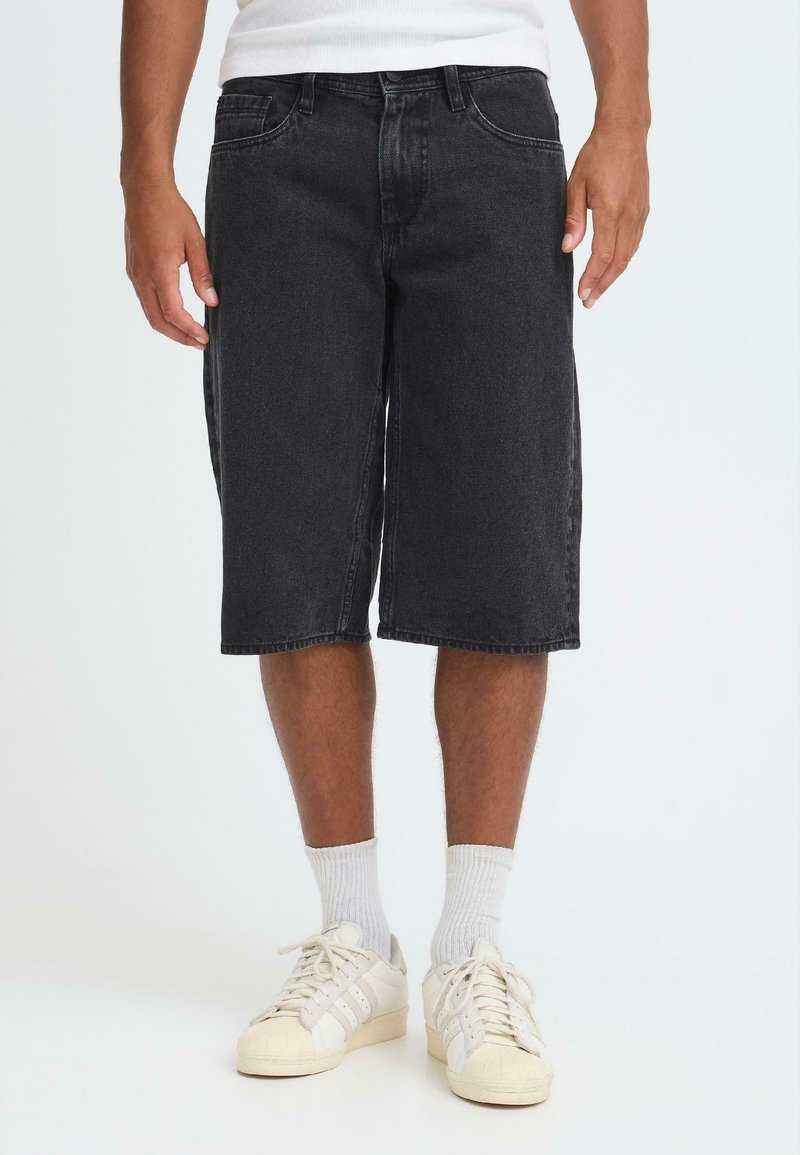 Shorts en denim noir à coupe droite, ourlet longueur genou, avec un design classique à cinq poches. Portés avec des chaussettes blanches et des baskets crème.