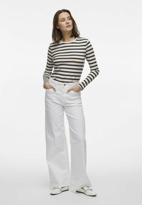 Randig långärmad topp i beige och marinblå, kombinerad med helvita högmidjade vida byxor. Modellen står med händerna i fickorna och vänder sig åt vänster.