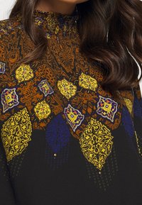 Top in tessuto nero con intricati motivi arancioni, gialli e blu, con colletto alto e dettagli decorativi, tra cui piccoli elementi metallici.