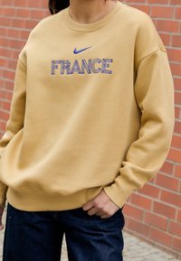 Osoba w beżowej bluzie z napisem "FRANCJA" oraz niebieskim logo Nike, w połączeniu z ciemnymi dżinsami, stojąca przy ceglanej ścianie.