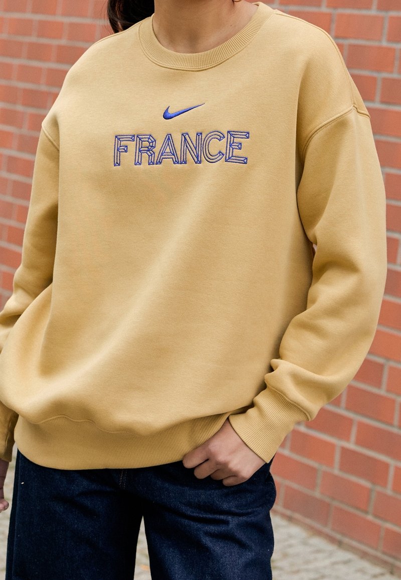 Osoba w beżowej bluzie z napisem "FRANCJA" oraz niebieskim logo Nike, w połączeniu z ciemnymi dżinsami, stojąca przy ceglanej ścianie.