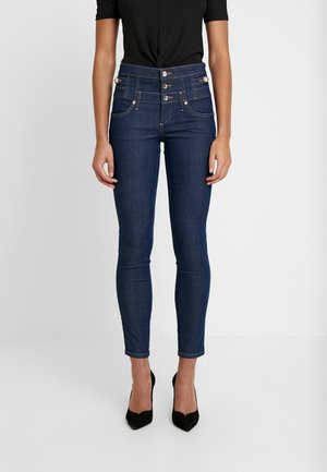 Frau trägt dunkelblaue High-Waist Skinny Jeans mit dreifachem Knopfverschluss und schwarzen spitzen High Heels, stehend vor einfarbigem Hintergrund.
