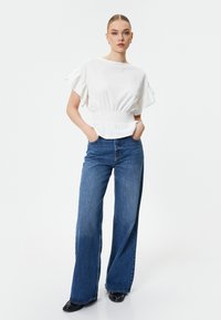 Hvid bluse med korte volangerede ærmer og smocket talje, parret med høj talje, brede blå jeans og sorte stiletter.