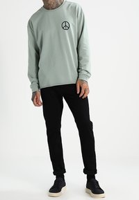 Felpa a maniche lunghe verde chiaro con simbolo della pace nero, abbinata a pantaloni neri e sneaker nere. Design semplice e casual.