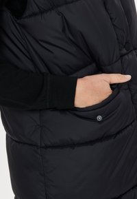 Svart pufferväst med vadderad design, med en sidoficka med tryckknappsstängning. Mjuk textur och rund halsringning synlig på bilden.