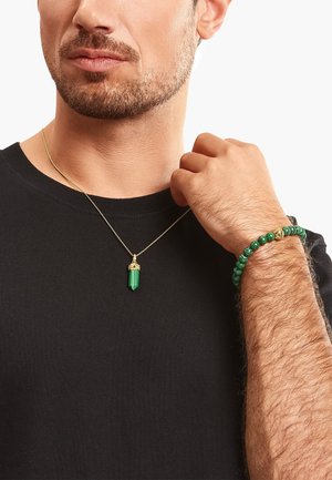 Una collana di catena d'oro con un pendente in cristallo verde; al polso c'è un braccialetto di perline verdi con dettagli dorati; indossato sopra una maglietta nera.