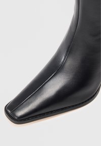 Bottine en cuir noir avec un bout pointu, présentant une texture lisse et des coutures minimales. La semelle est d'une couleur claire contrastante.