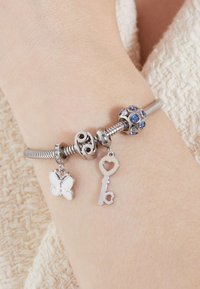 Silberne Charm-Armbanduhr mit einem weißen Schmetterlingscharme, einem herzförmigen Schlüssel und einer blau akzentuierten Perle, ausgestattet mit einem flexiblen Metallkettendesign.