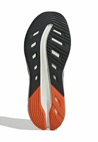 La semelle de la chaussure présente une surface noire texturée avec des accents blancs et une section orange. Des motifs rainurés améliorent l'adhérence et la traction.