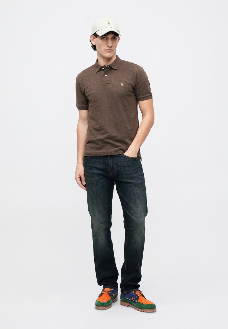 Junger Mann steht da und trägt ein braunes Poloshirt, dunkle Jeans, mehrfarbige Schuhe und eine weiße Baseballmütze vor einem schlichten weißen Hintergrund.