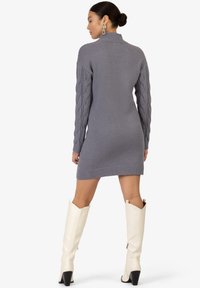 Abito maglione grigio con un colletto alto, caratterizzato da maniche a treccia, polsini a coste e lunghezza fino a metà coscia. Indossato con stivali bianchi fino al ginocchio.
