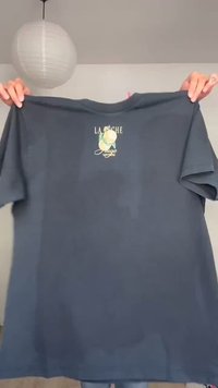 T-shirt gris foncé tenu par des mains avec un petit motif de citron à l'avant et le texte "LA POCHE" et "Lugo" sur un fond uni.