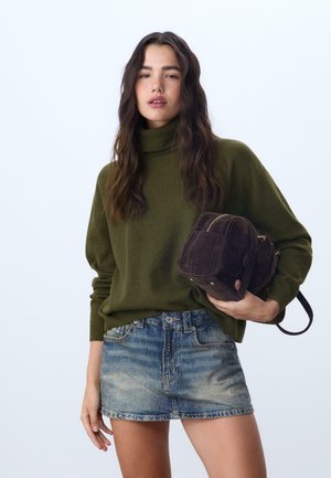 Frau mit langen dunklen Haaren, die einen olivgrünen Rollkragenpullover, einen Jeans-Minirock trägt und einen dunkelbraunen Cordura-Rucksack hält.