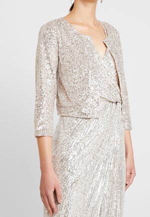 Kvinde iført en sølv paliet cropped jakke over en matchende ærmeløs V-hals kjole, stående mod en ensfarvet lys baggrund.