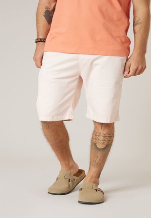 Homme portant un short rose clair, un t-shirt saumon, des chaussures beige sans lacets, et ayant des tatouages visibles sur les jambes et les bras, debout sur un fond uni.