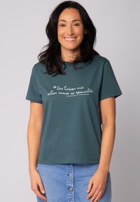 Teal katoenen T-shirt met korte mouwen, voorzien van witte tekst in een casual lettertype op de voorkant. Draagt met een spijkerrok, wat een ontspannen pasvorm laat zien.