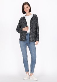 Jachetă neagră cu model floral, cu fermoar în față, din material ușor, și cu glugă, purtată cu blugi skinny de un albastru deschis și pantofi albi.