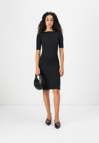 Woman with curly hair wearing a fitted black knee-length dress, carrying a black handbag, standing against a plain white background.