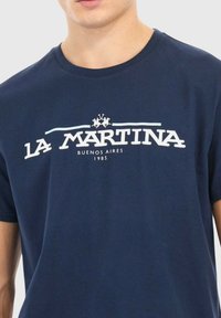 Man som bär en marinblå T-shirt med vitt "La Martina Buenos Aires 1985"-logotyp och två polospelare grafik på bröstet.