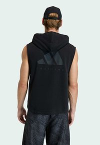 Schwarzer ärmelloser Hoodie mit großem grafischem Logo auf dem Rücken, kombiniert mit dunklen gemusterten Shorts. Der Hoodie verfügt über eine vordere Kängurutasche.